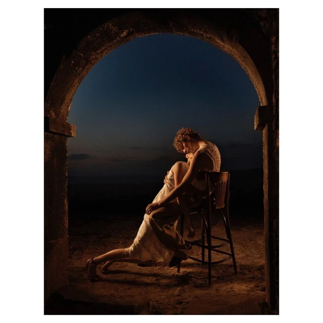 Intimate portrait of a couple in a stone archway at dusk, with a woman seated on a chair, embracing. Warm, romantic lighting.