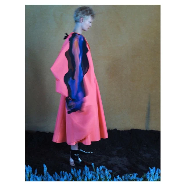 Fashion shot of a slender person in a pink coat with colorful accents, silver booties, and a neutral backdrop, merging art and fashion.