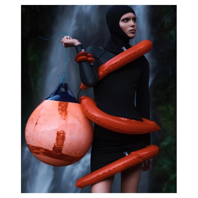 Model in black wetsuit and hood poses with buoys and inflatable armbands against a waterfall backdrop for fashion editorial.