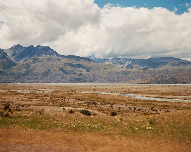 Scenic view of a valley with brown grass, a distant lake, and mountains beneath a partly cloudy sky, capturing nature's serene beauty.