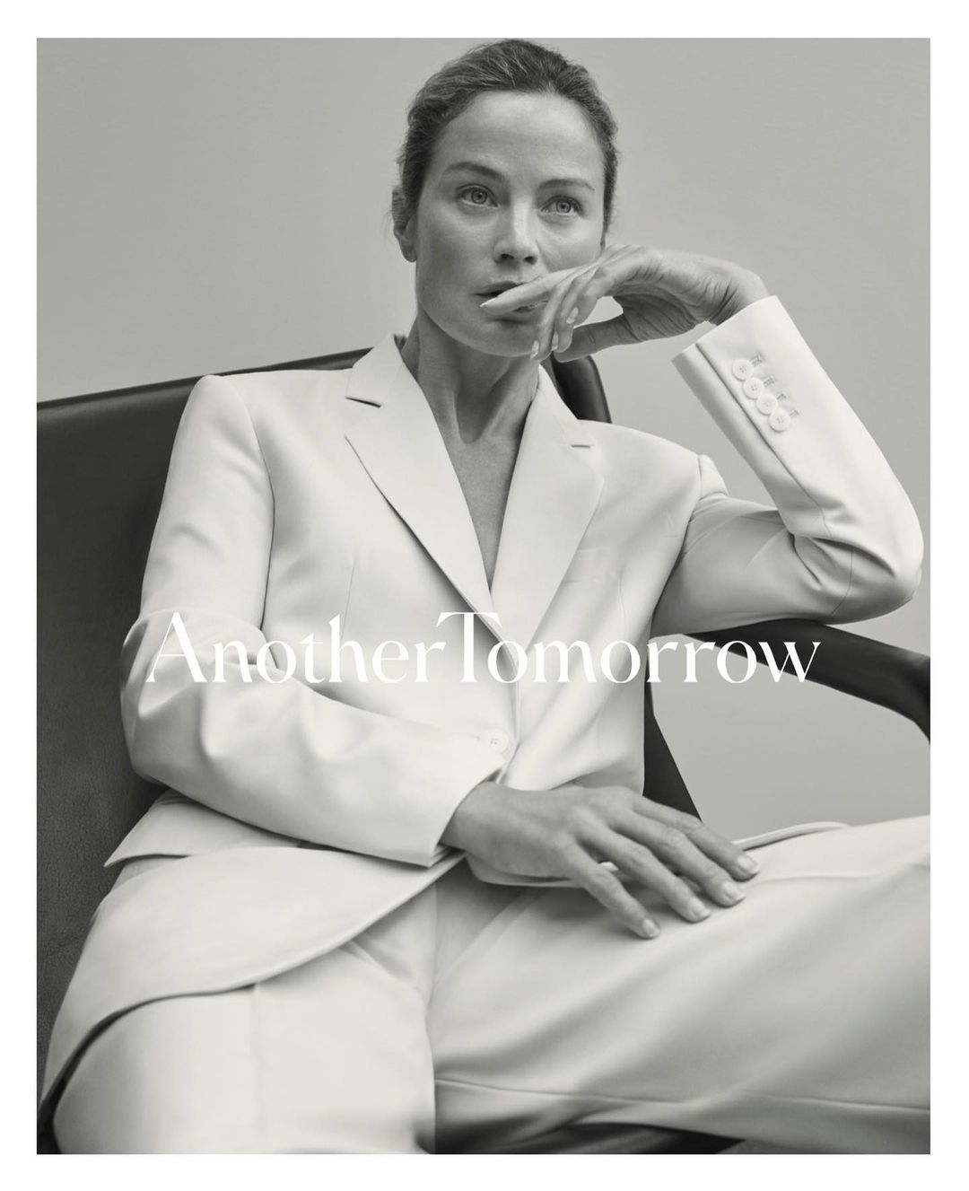 Black and white portrait of an adult woman with hand near her mouth in a chair, wearing a white suit on a gray background.