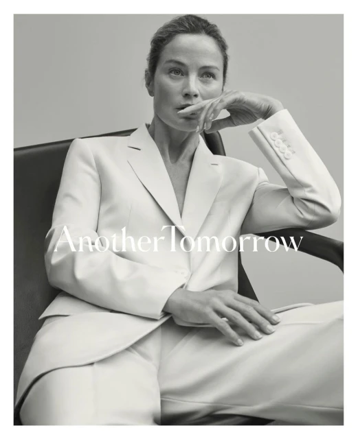 Black and white portrait of an adult woman with hand near her mouth in a chair, wearing a white suit on a gray background.