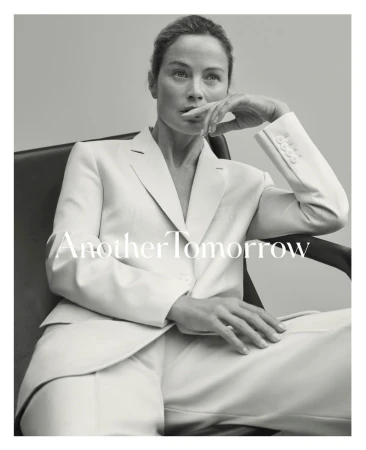 Black and white portrait of an adult woman with hand near her mouth in a chair, wearing a white suit on a gray background.