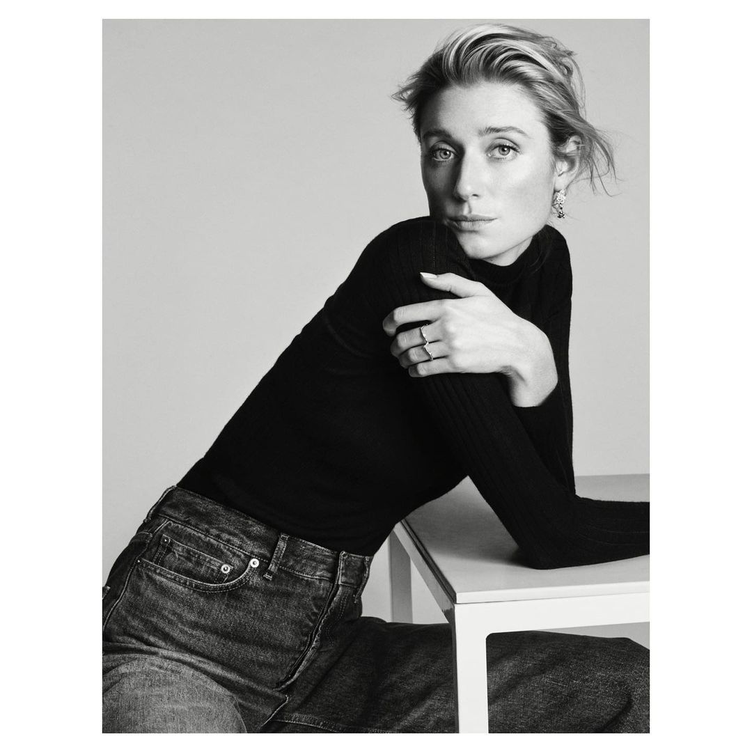 Black and white portrait of a woman in a black turtleneck and dark jeans leaning on a table by Josh Olins