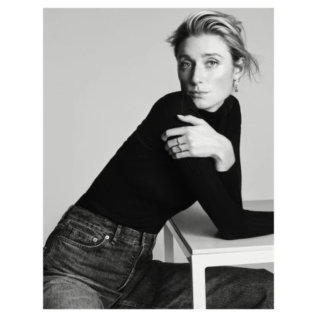 Black and white portrait of a woman in a black turtleneck and dark jeans leaning on a table by Josh Olins