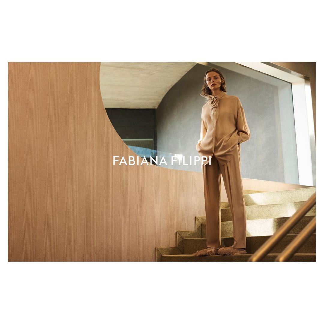 A woman in a tan sweater and pants stands on stairs next to a tan wall with an oval mirror in a fashion-style, high-key shot.