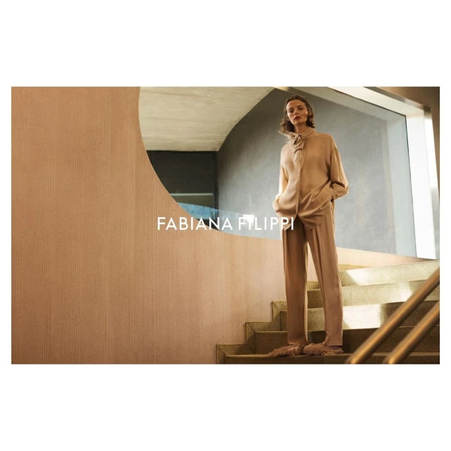 A woman in a tan sweater and pants stands on stairs next to a tan wall with an oval mirror in a fashion-style, high-key shot.