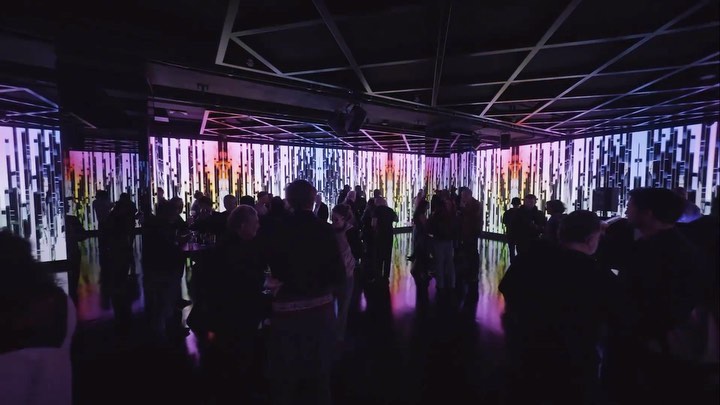 Large group of people gathered in a dark indoor space with colorful vertical lights and metal ceiling structure, creating an event atmosphere.