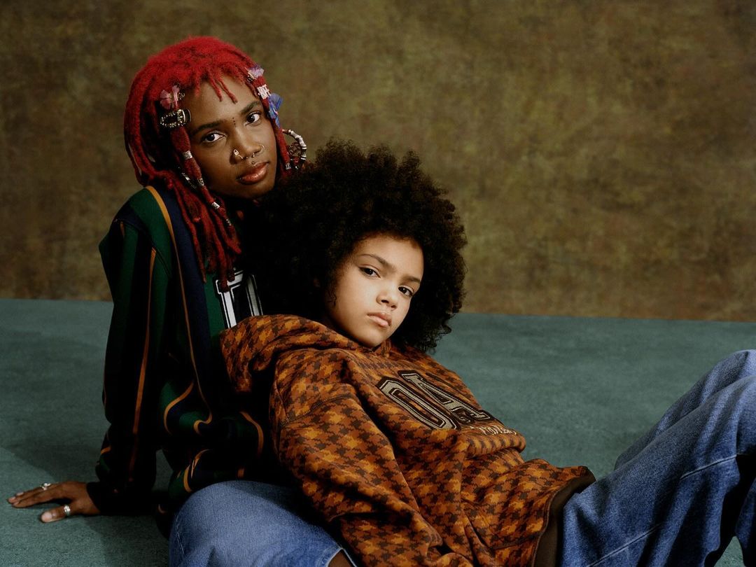 Two young people pose in an indoor studio setting, one with red dreadlocks, wearing a striped sweater, the other in a printed hoodie and jeans.