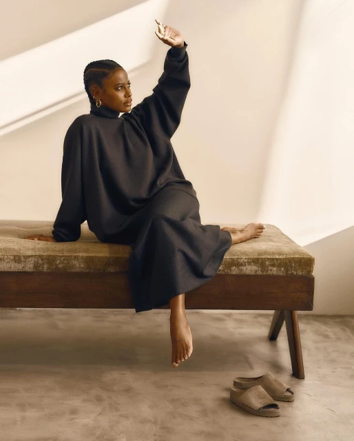 Woman in long black dress and hoop earrings sits on a velvet bench and raises hand, casting a shadow against a cream wall in studio.