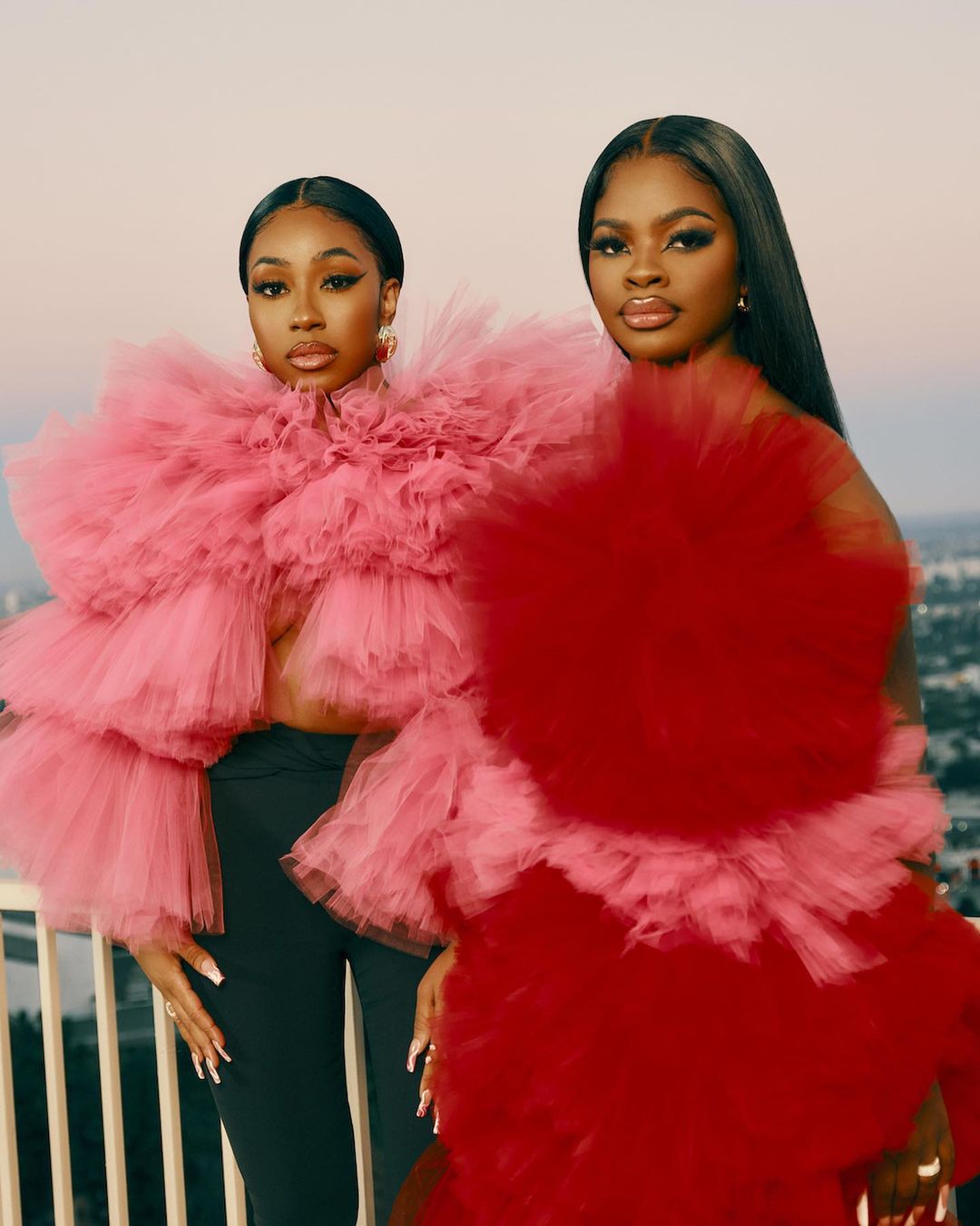 Two Black women pose in oversized pink and red ruffled tops against an outdoor cityscape backdrop, presenting a striking fashion portrait.