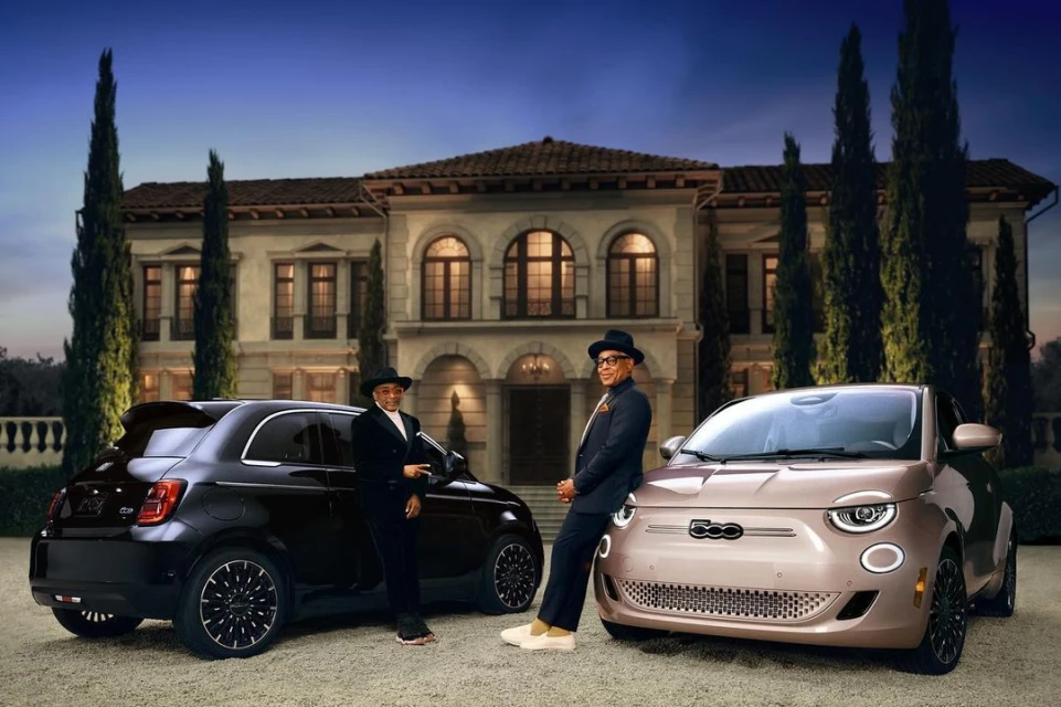 Two individuals stand beside a dark Fiat 500e and a beige Fiat 500e in front of a villa, showcasing automotive elegance and style.