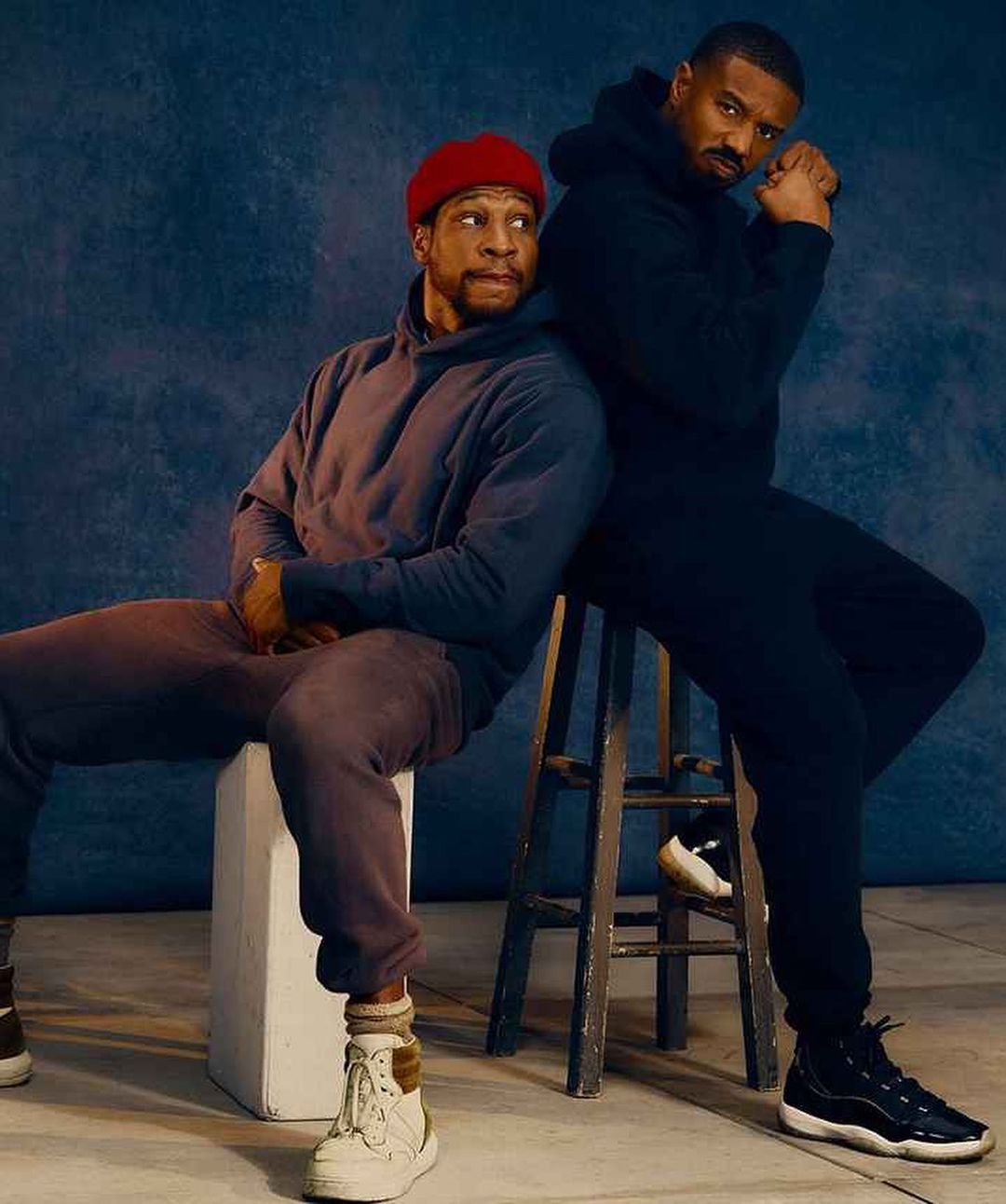 Two adult males in dark sweatsuits sitting indoors. One man leans against the other who is seated on a stool, in a studio setting.