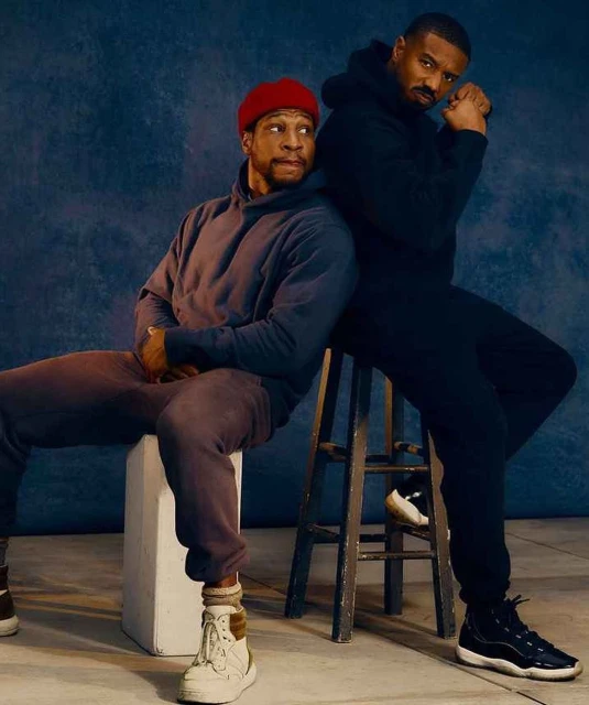 Two adult males in dark sweatsuits sitting indoors. One man leans against the other who is seated on a stool, in a studio setting.