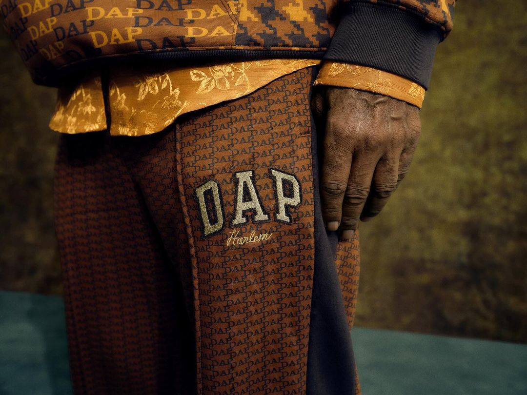Close-up of a person's torso and hand wearing a DAP-branded tracksuit. Garments feature a brown, tan, and black color palette.