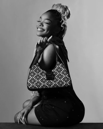Black and white studio shot of smiling woman holding purse with floral pattern, wearing dark dress and hair styled in a high bun.