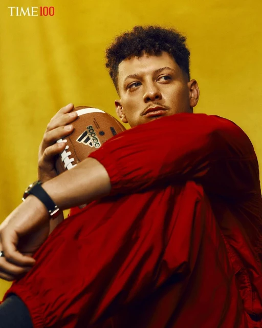 Portrait of football player in red jacket holding Adidas football against yellow background, wearing black wristwatch and rings. 