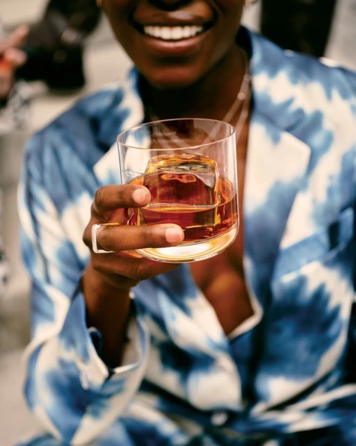 Smiling woman in blue tie-dye robe holds clear glass of amber liquor with large ice cube, accessorized with ring and necklace.
