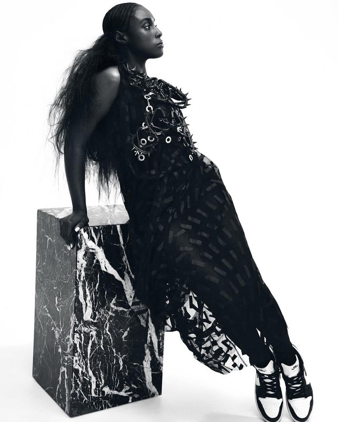 An adult woman in a dress and sneakers leans against a marble cube in a studio, looking to the right in this black and white shot.