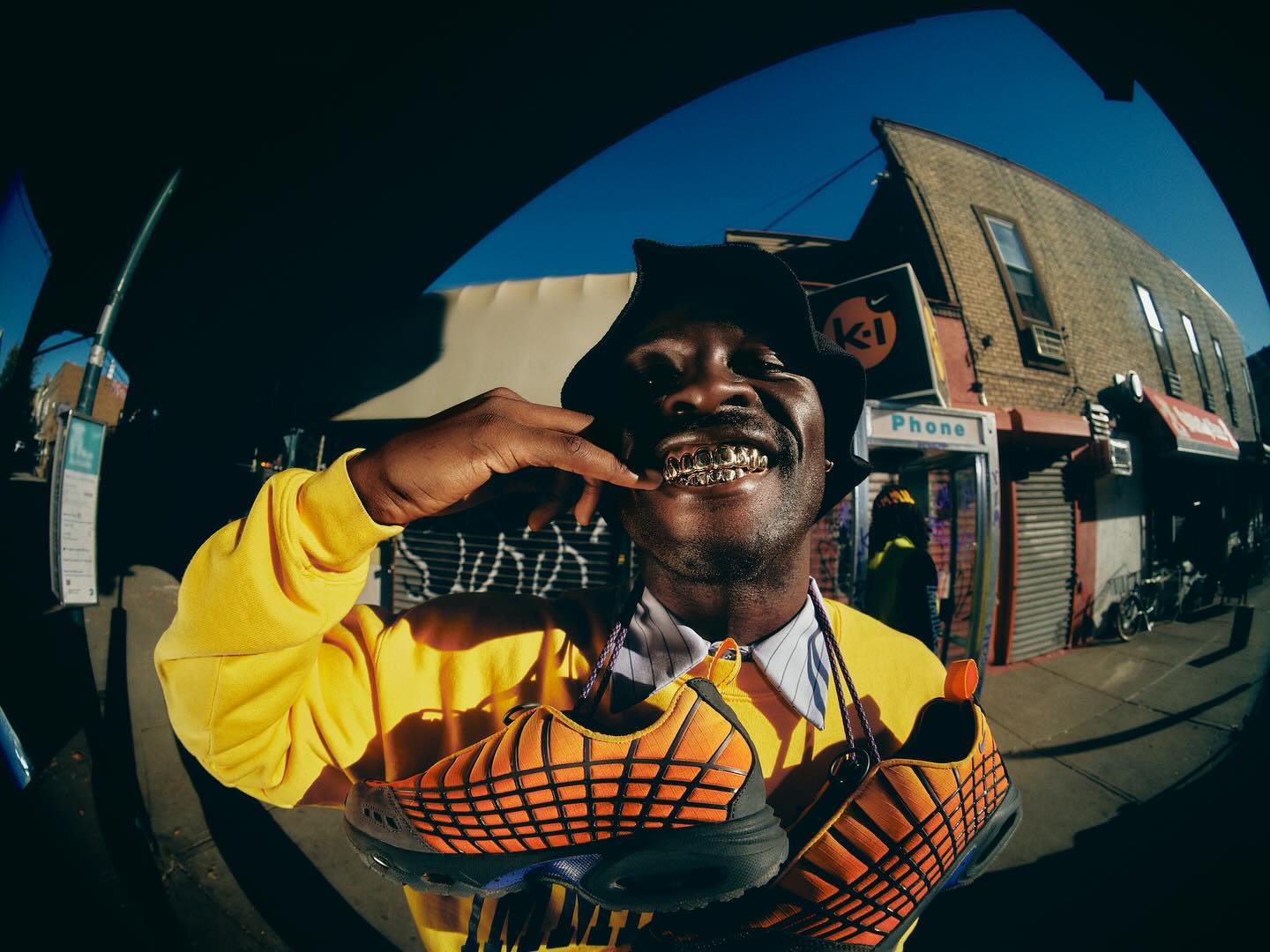 Man wearing bucket hat and yellow sweatshirt smiles holding orange and black athletic shoes in an urban setting on a sunny day.
