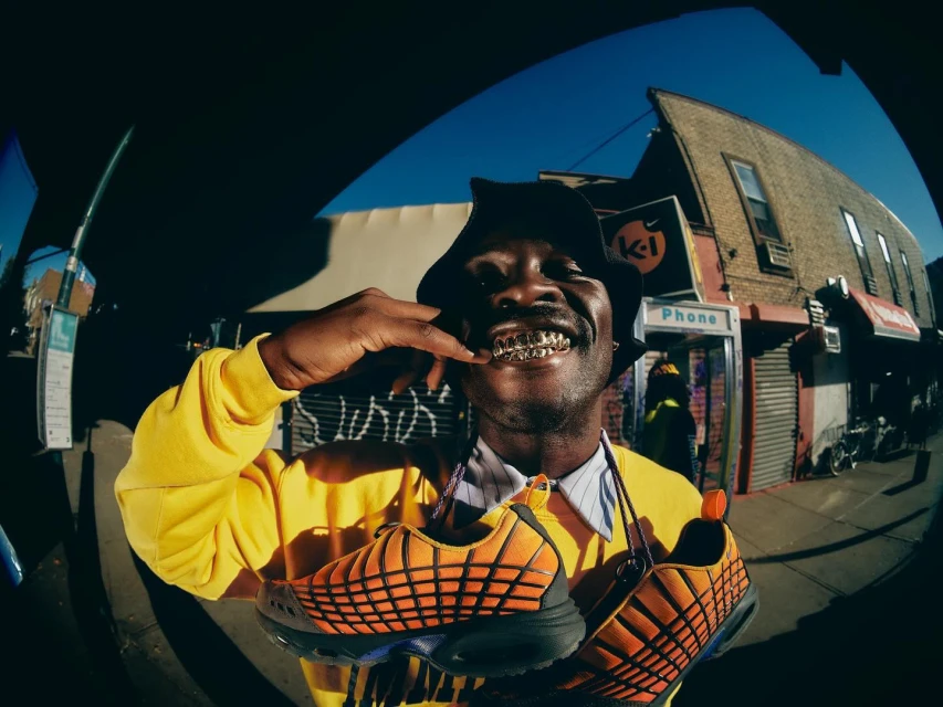 Man wearing bucket hat and yellow sweatshirt smiles holding orange and black athletic shoes in an urban setting on a sunny day.