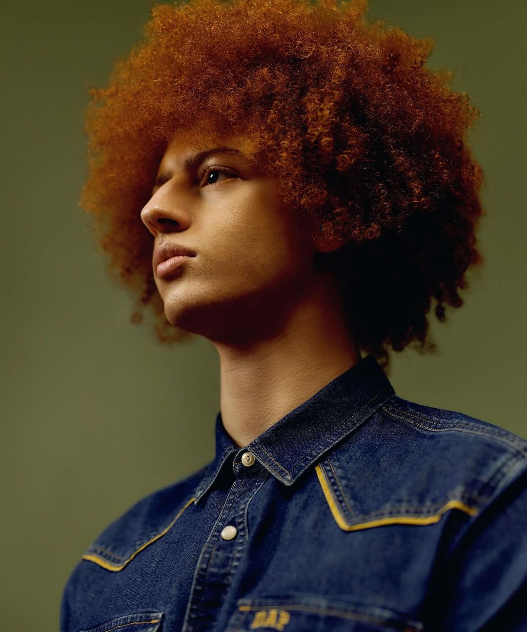 Portrait of a young man with an afro wearing a denim shirt, set against an olive-green background in a studio setting, looking off-camera.