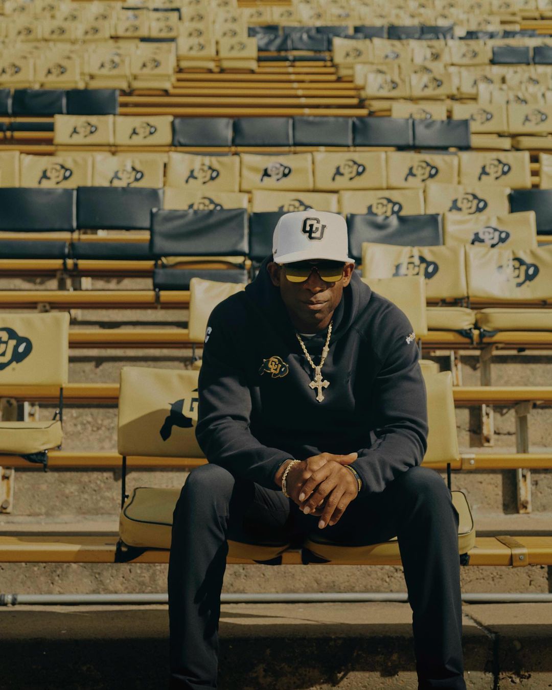 Football coach Deion Sanders is seen in a medium shot, sitting in stadium seating wearing team apparel and a baseball cap.
