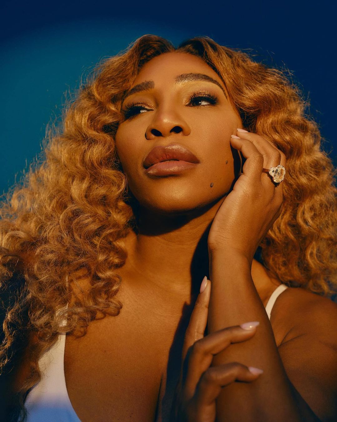 Close-up of Serena Williams with golden curly hair, hand on her face, wearing a white top against a blue backdrop, ring visible.
