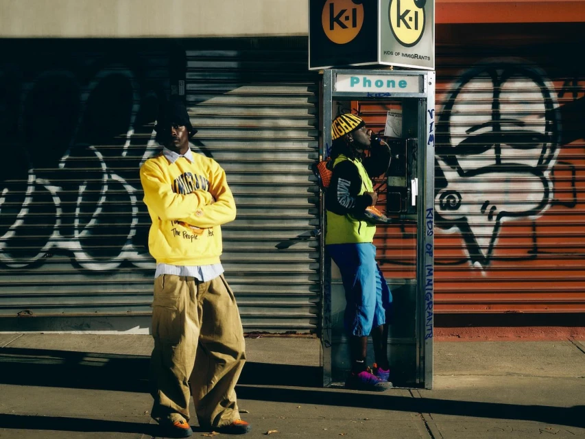 Two men in street style fashion pose in an urban setting. One uses a phone booth in neon colors and graffiti for a fashion editorial.