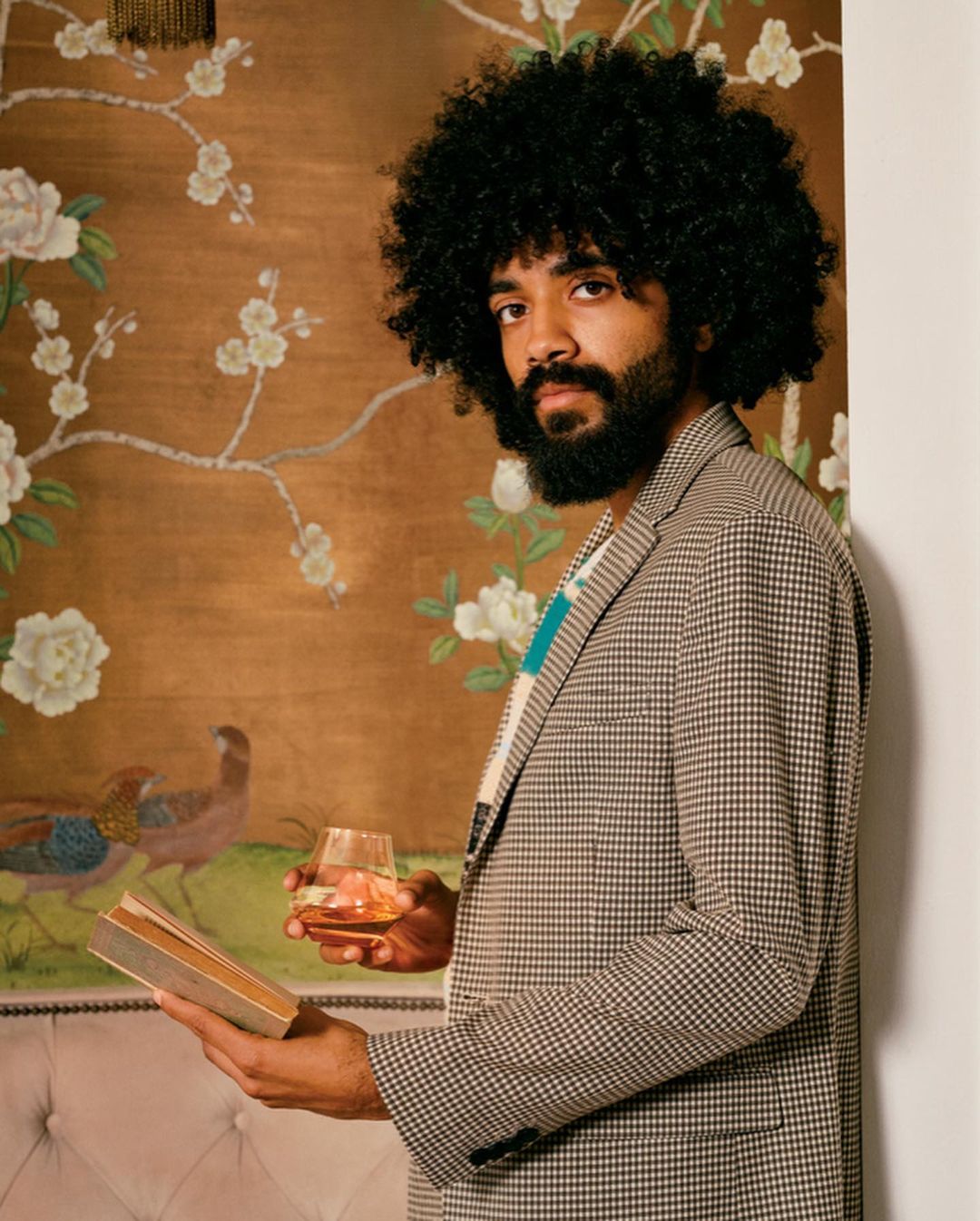 Man in houndstooth blazer holds book and whiskey glass, posing near floral art. He has an afro and beard, indoor setting, medium shot.