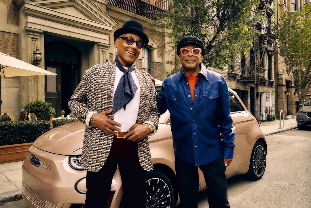 Two men stand beside a bronze-colored FIAT 500 car on a city street, smiling and wearing stylish clothing on a sunny day.