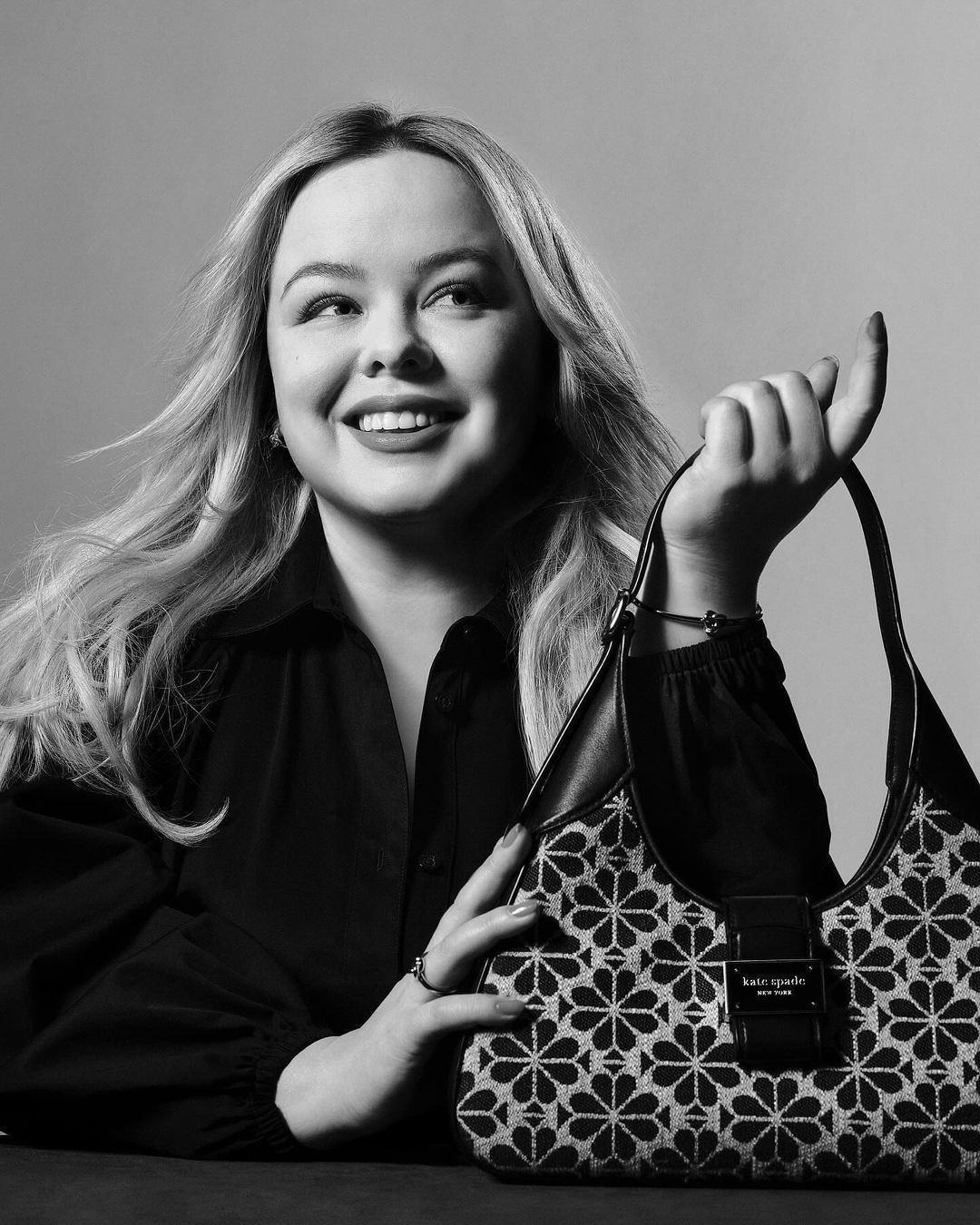 Monochrome portrait of a blonde woman smiling and holding a patterned black and white handbag in a studio setting with soft lighting.