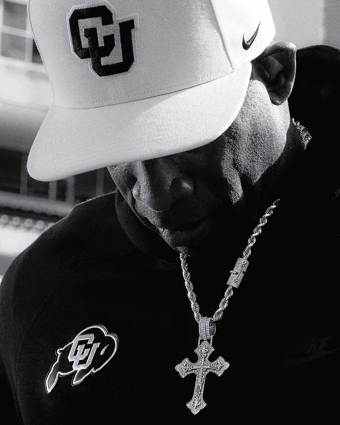 Monochrome portrait of an adult male wearing a branded cap, dark hooded sweatshirt, and a prominent cross pendant necklace. 