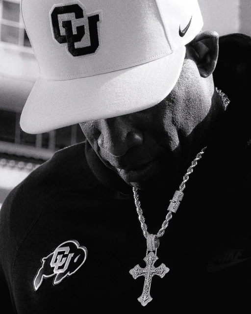 Monochrome portrait of an adult male wearing a branded cap, dark hooded sweatshirt, and a prominent cross pendant necklace. 