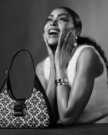 Black and white portrait of a woman laughing with her hands on her cheeks, beside a patterned shoulder bag.