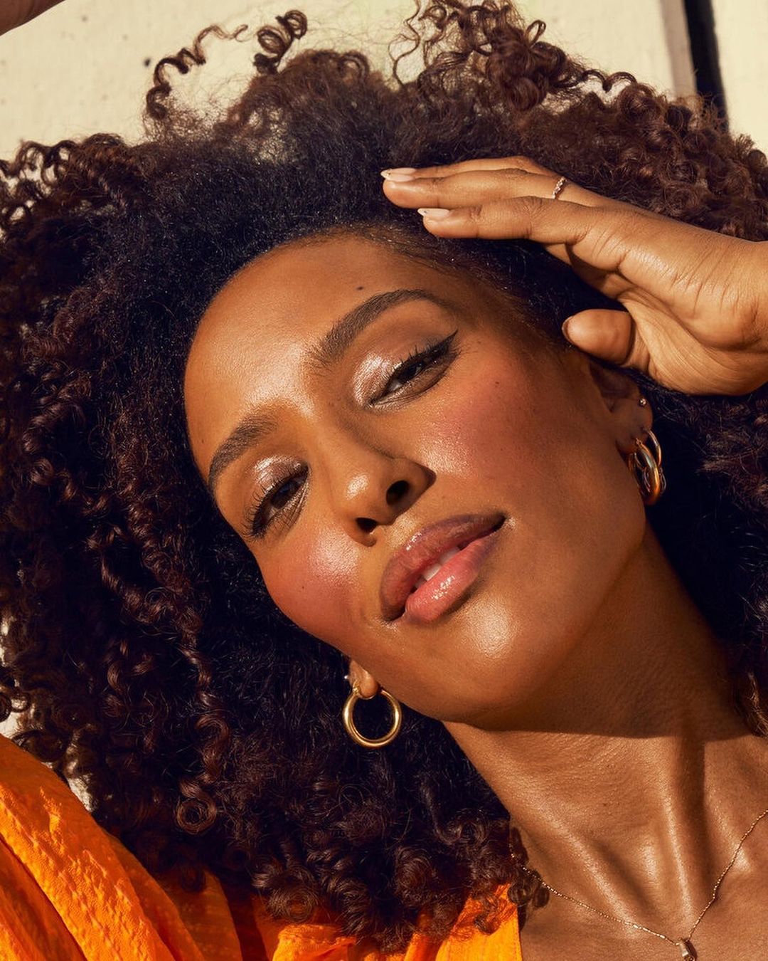 Close-up of a woman with dark curly hair and tan skin wearing gold hoop earrings and an orange top against a neutral background.