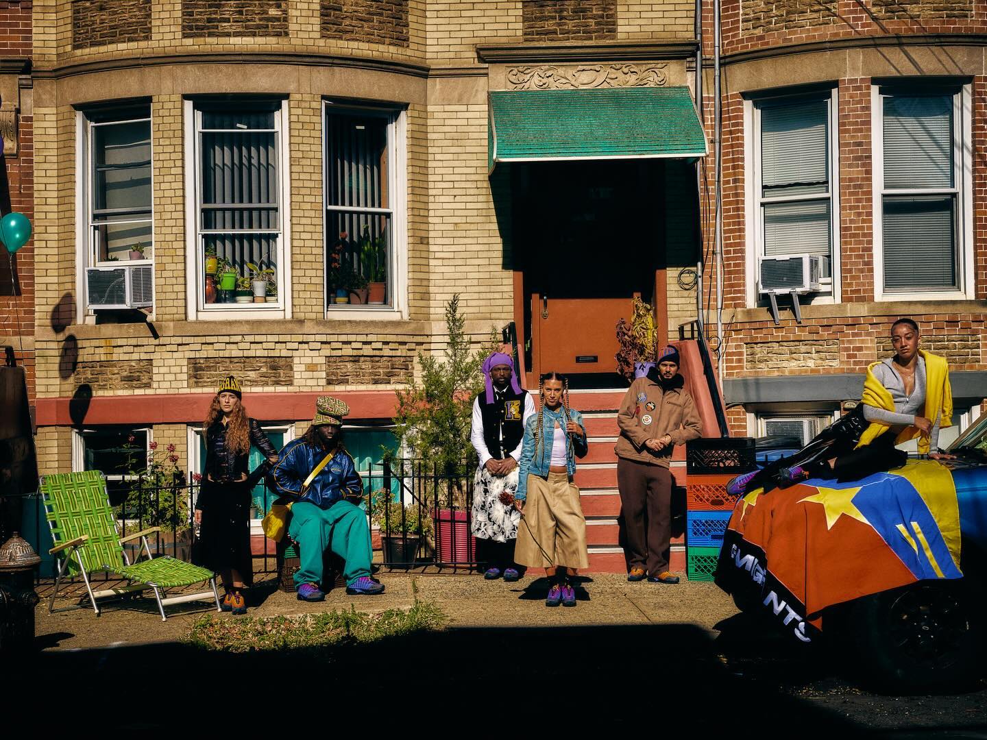 Six adults pose in front of a brick apartment building with green awning in daylight. A car with a colorful blanket and balloon visible.