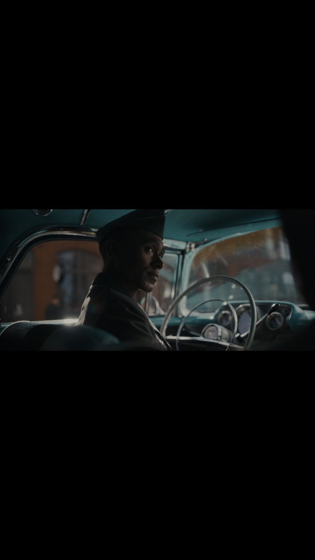 Adult male driving vintage car in urban setting. He is wearing a military-style cap and looks toward the camera from inside the car.