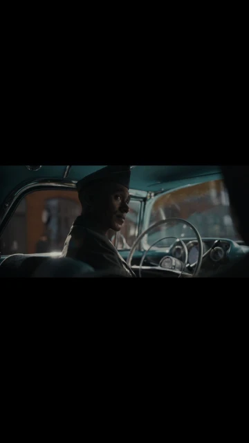 Adult male driving vintage car in urban setting. He is wearing a military-style cap and looks toward the camera from inside the car.