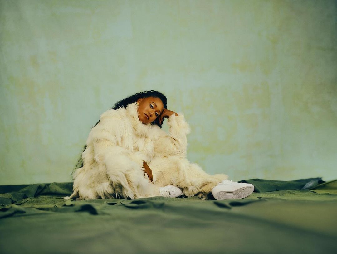 Woman with dark hair posing in fluffy white jacket and sneakers, seated on green fabric against a light green backdrop in a studio setting.
