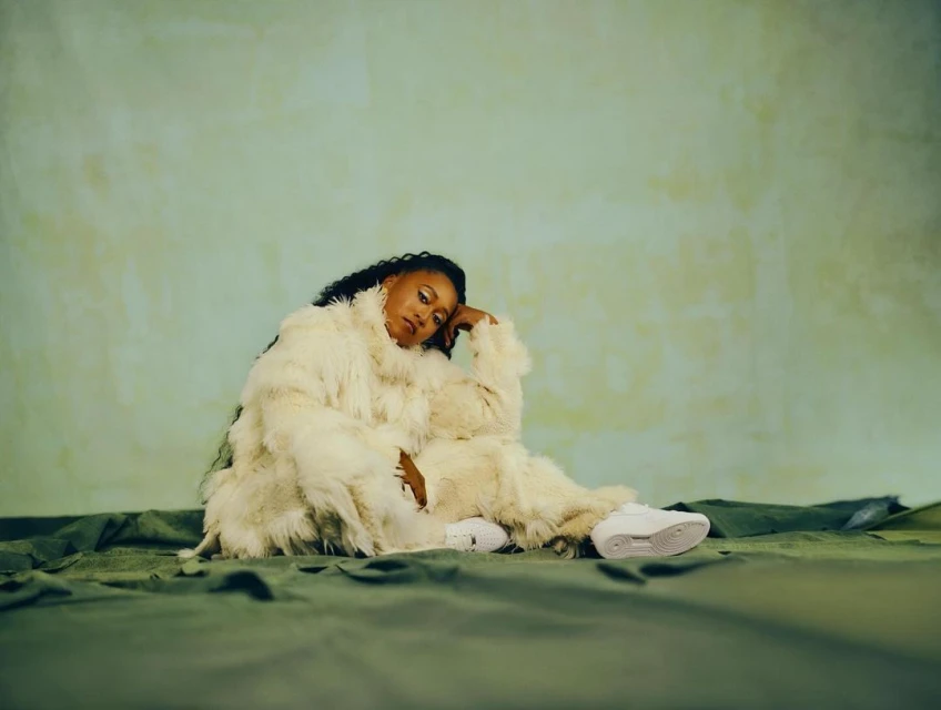Woman with dark hair posing in fluffy white jacket and sneakers, seated on green fabric against a light green backdrop in a studio setting.