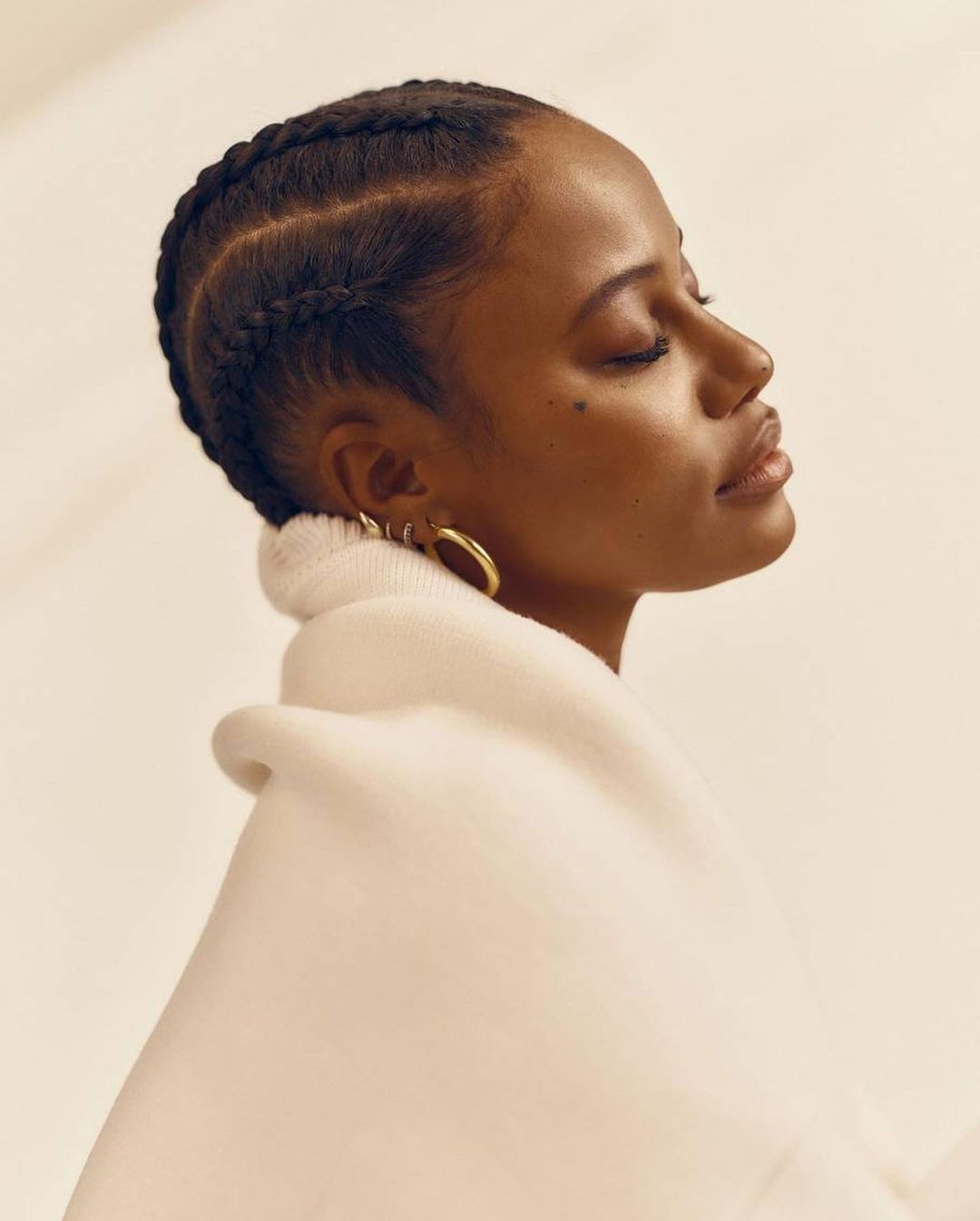 Profile portrait of a Black woman with braided hair wearing gold hoop earrings and wrapped in a cream textile against a neutral backdrop.