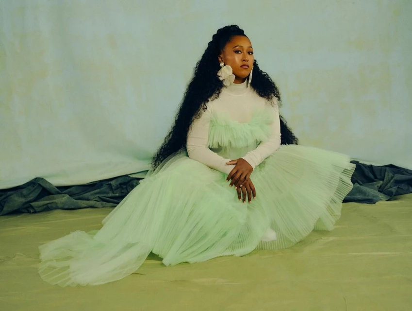 Adult female seated wearing a white turtleneck top with a light green dress, large floral earrings and long dark curly hair in an indoor setting.