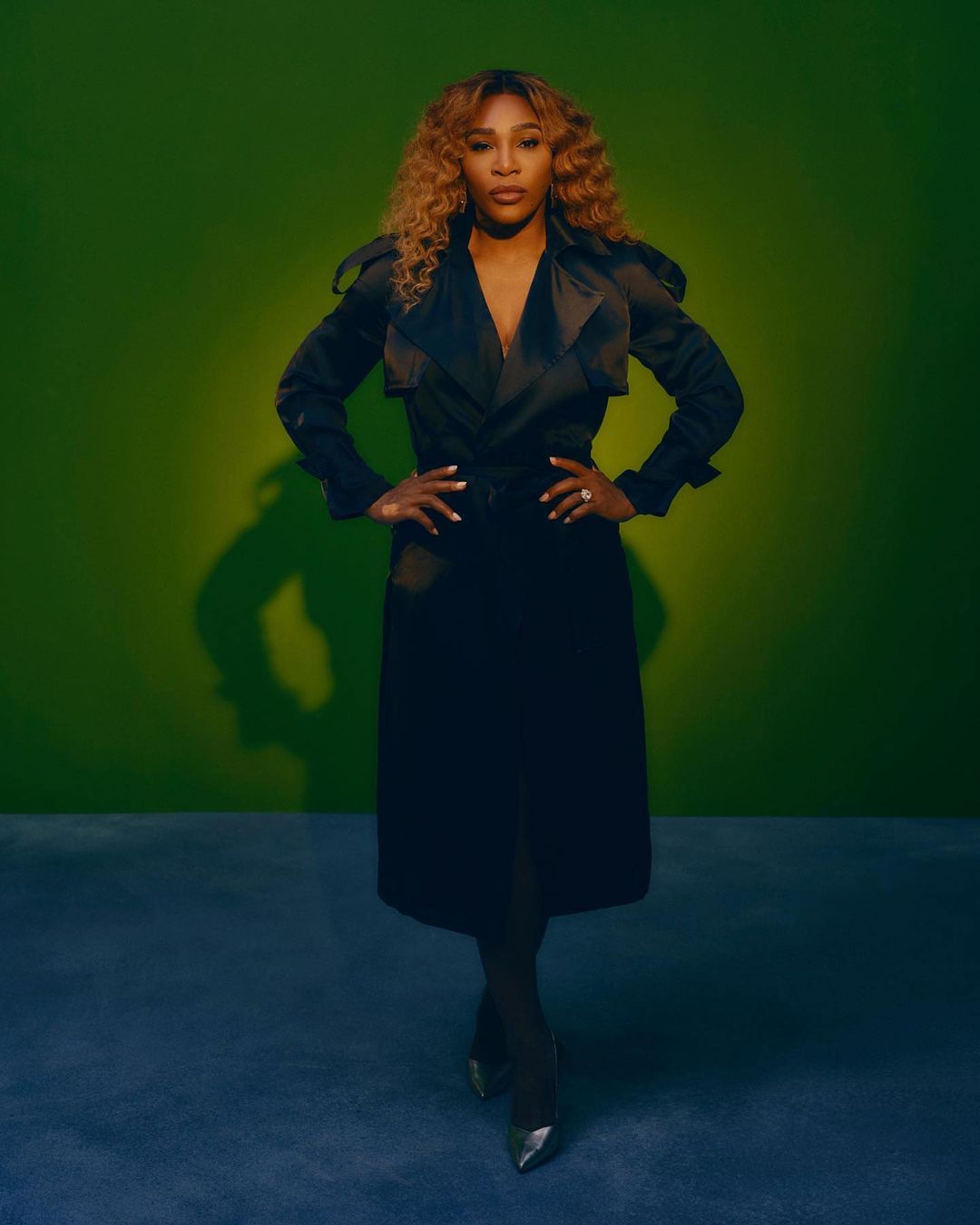 Serena Williams poses in a black trench coat against a green backdrop with studio lighting, showcasing an elegant and confident full-body shot.