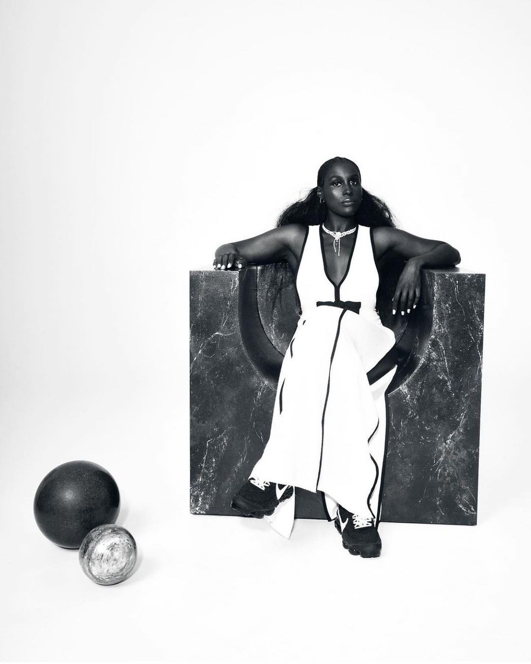 A fashionable dark-skinned adult female model sits next to sphere objects in a studio wearing a white dress in a portrait shot.