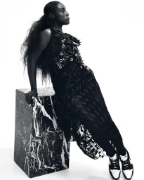 Black woman wearing a dark patterned dress and sneakers leaning against a marble block in a studio portrait