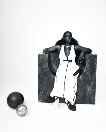 Black woman seated in a large dark sculptural chair wearing a white dress and black sneakers photographed by Joshua Kissi.