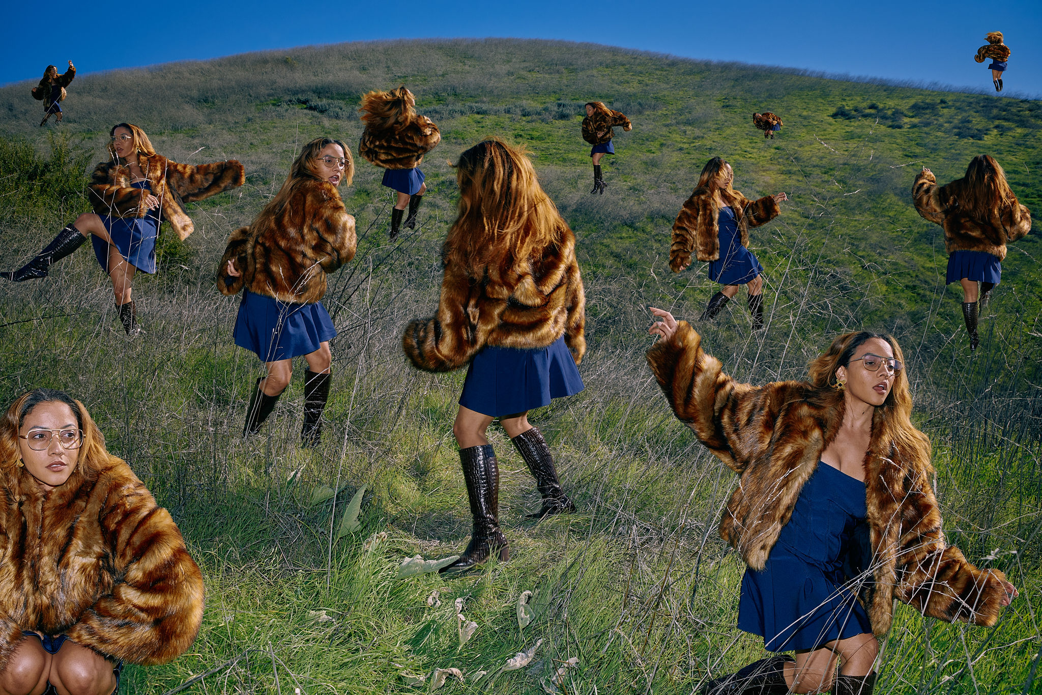 Several identical young women in fur coats pose in a grassy field under a blue sky, creating a surreal, dreamlike fashion scene.