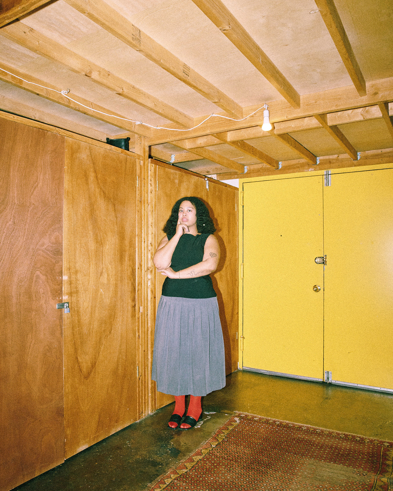Woman in a black top and grey skirt stands in an indoor room with yellow and wooden paneled walls and a patterned brown rug on the floor.