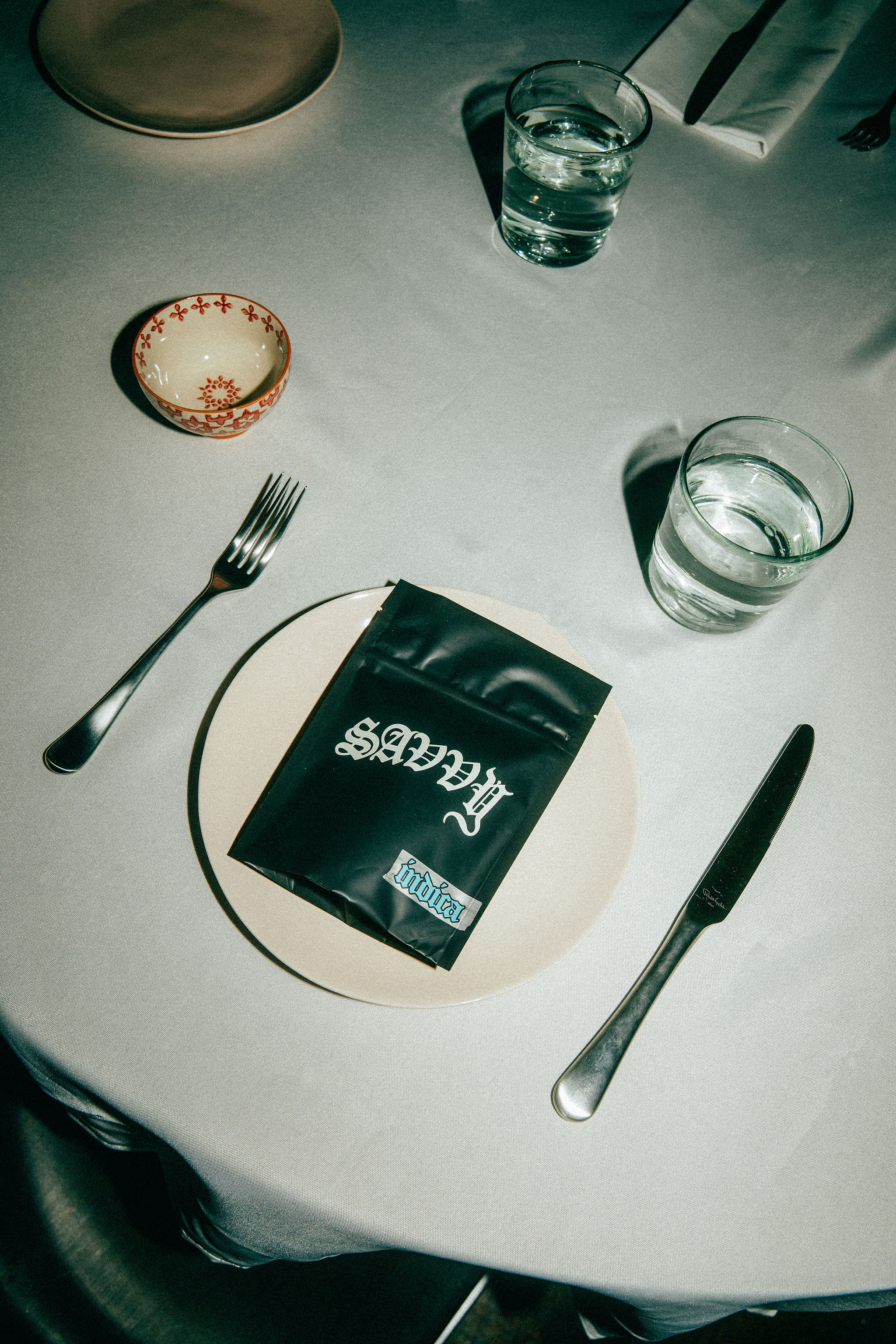 Overhead shot of black 'Savvy' product bag on white plate, surrounded by silverware, drinking glasses, and tablecloth.